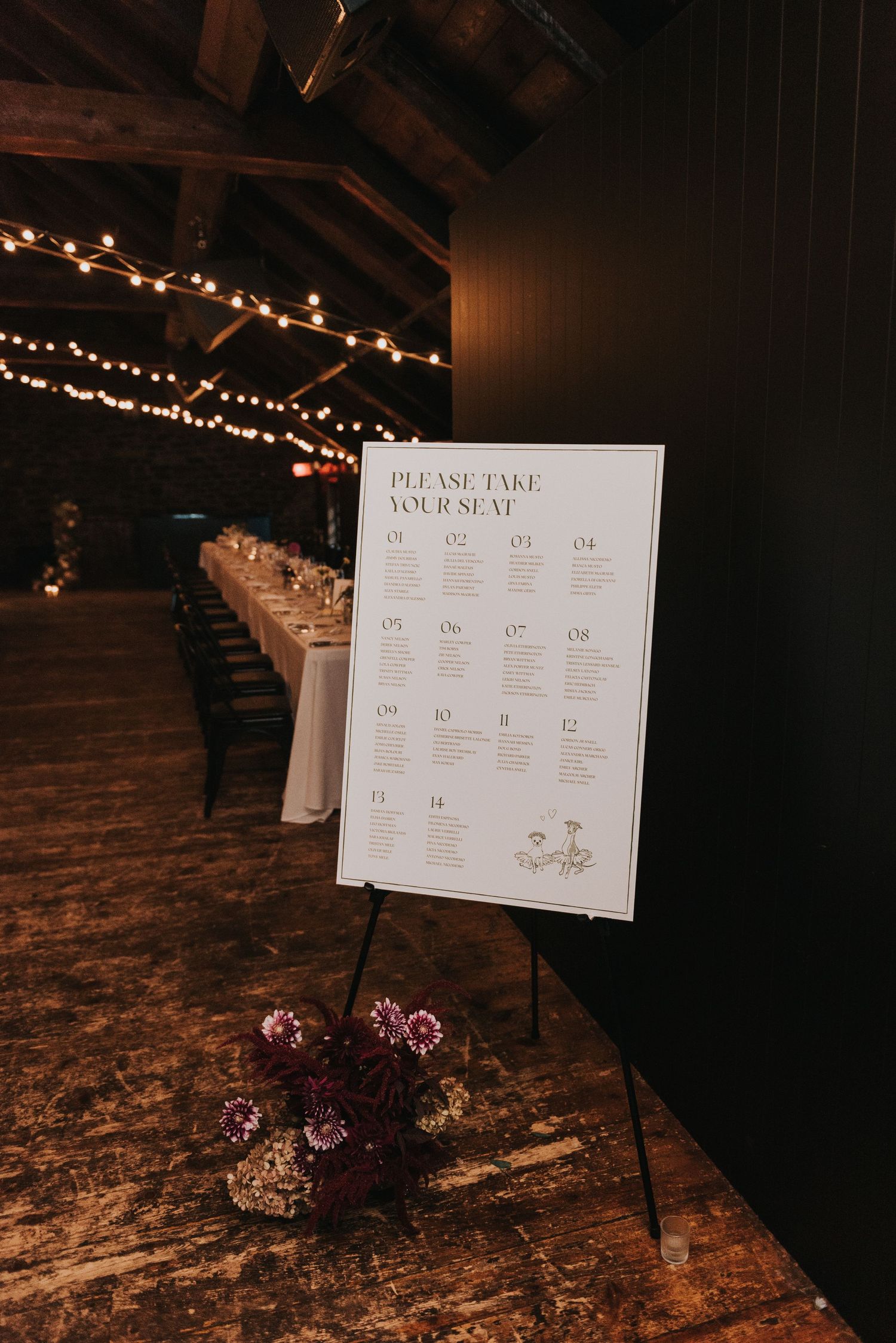 Wedding seating chart displayed on easel with string lights and purple flowers in rustic wooden venue.