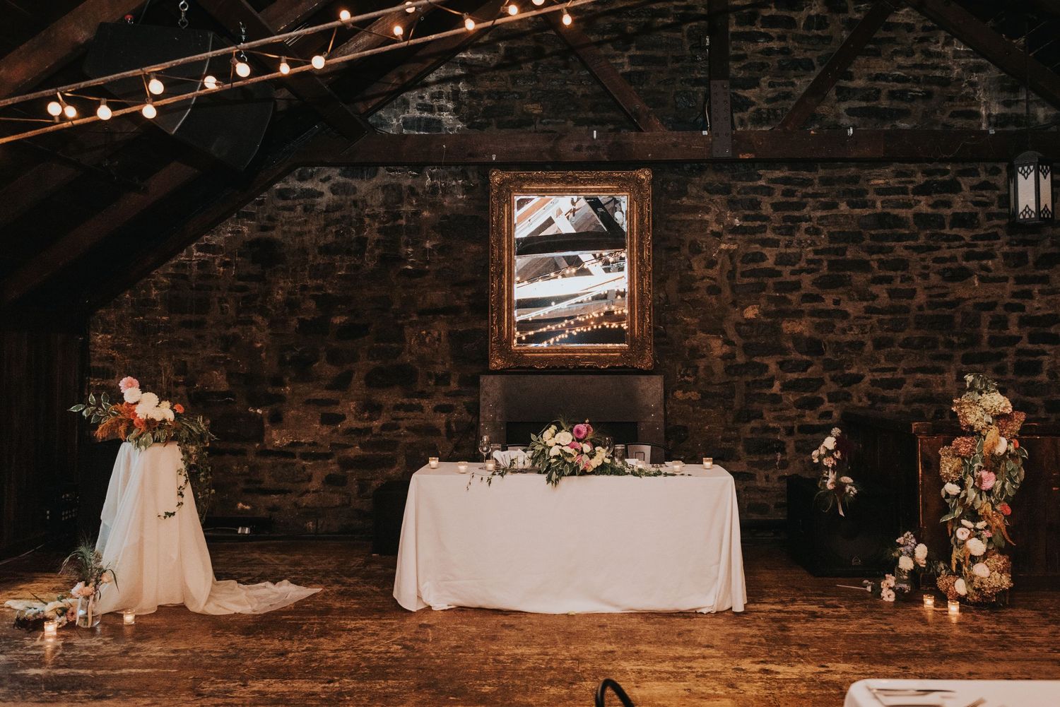 Rustic stone barn wedding venue interior with string lights, white tablecloth, and floral decorations.