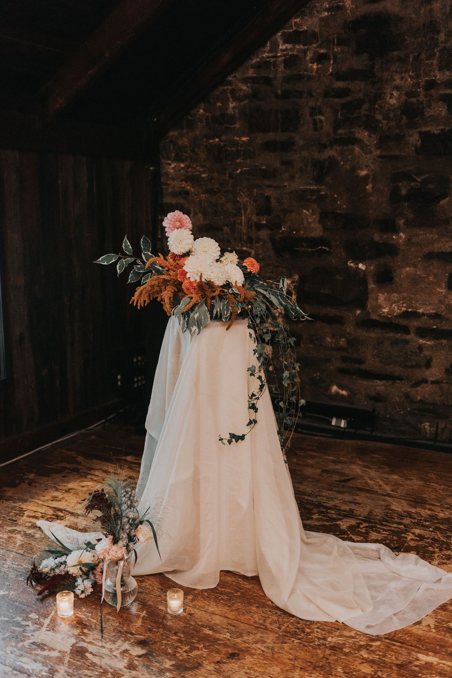A romantic floral arrangement with white and orange blooms cascades over white fabric against rustic brick walls.