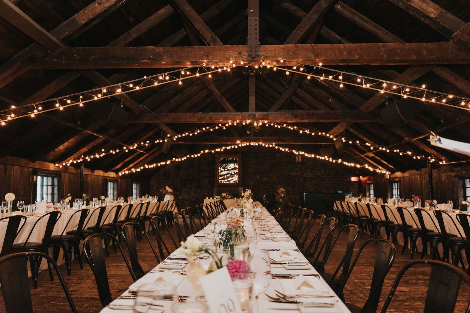 Long rustic wedding reception table set under wooden beams with string lights in a barn venue.
