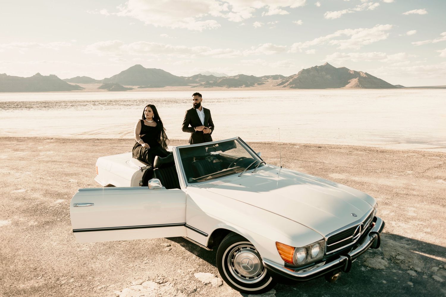 A classic silver Mercedes-Benz convertible parked on salt flats with mountains in the background at sunset.