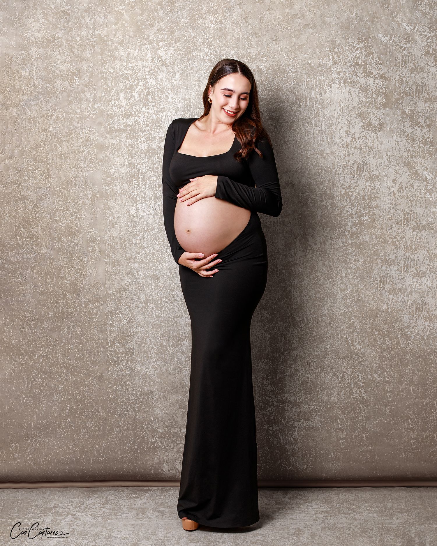 Mum in a modern black dress, hands gently on her bump, in a classic studio portrait.