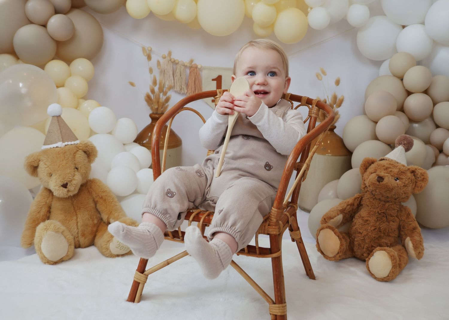 Baby in white dress sitting with gold and white birthday cake against balloon backdrop. Coventry Cake Smash Photographer. First Birthday.