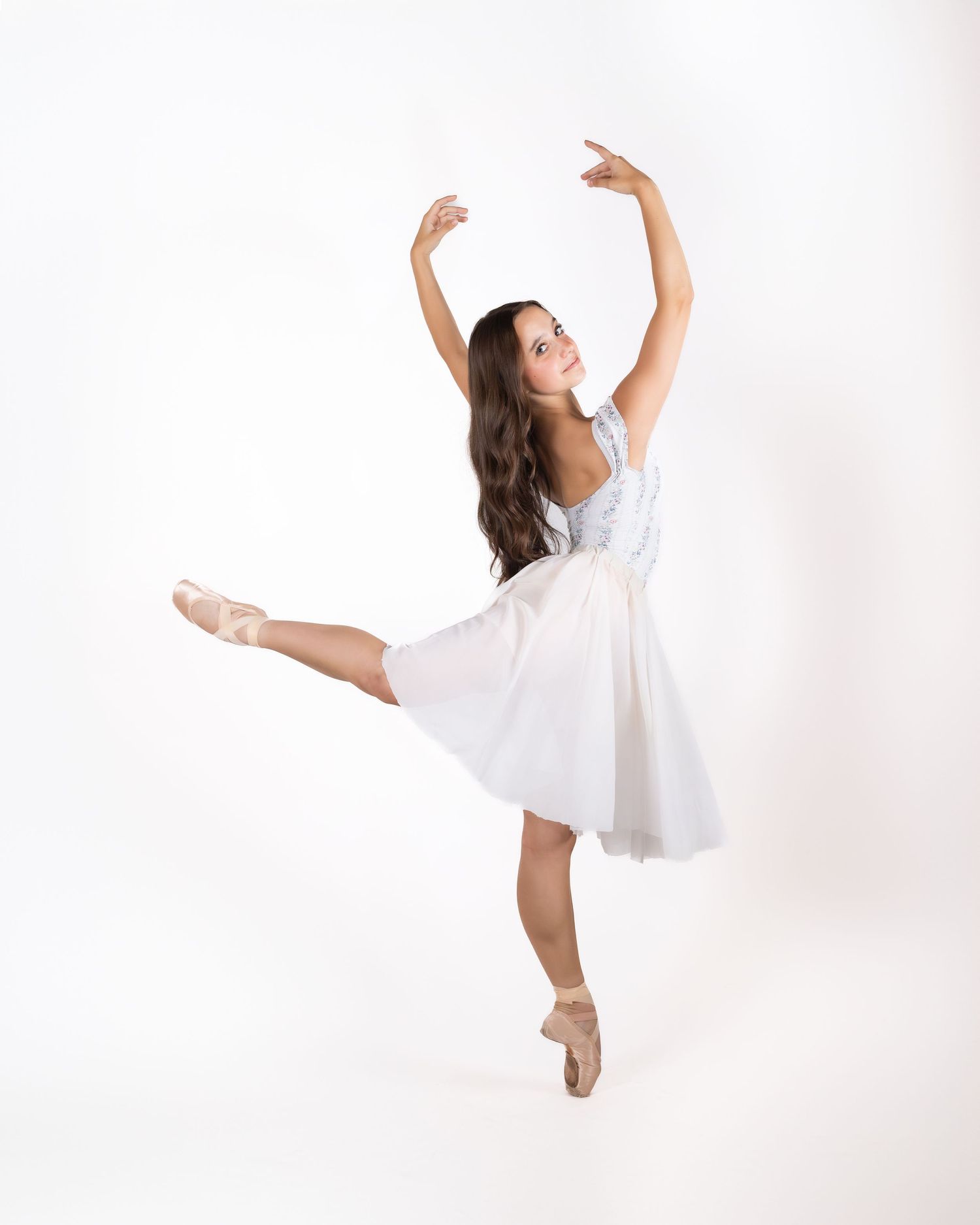A ballet dancer in a flowing white dress performs an elegant pose on pointe shoes against a light background.