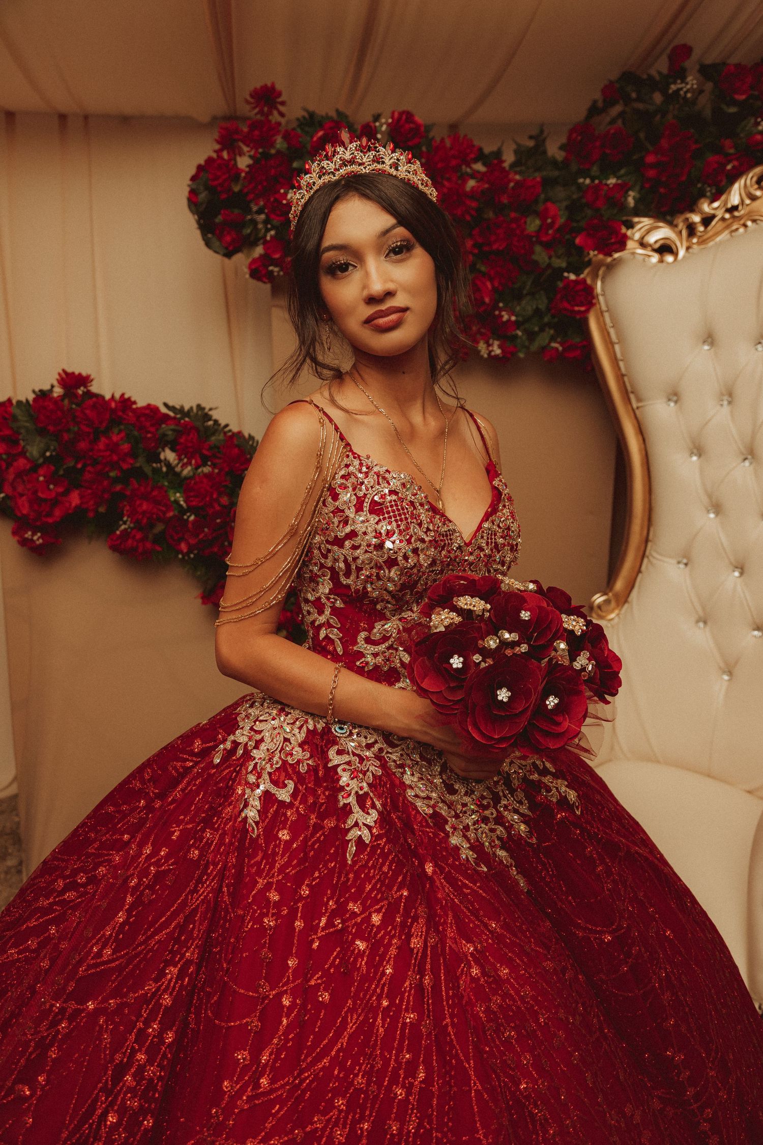 A model in a luxurious red and silver sequined ball gown sits on a cream tufted chair surrounded by red roses.