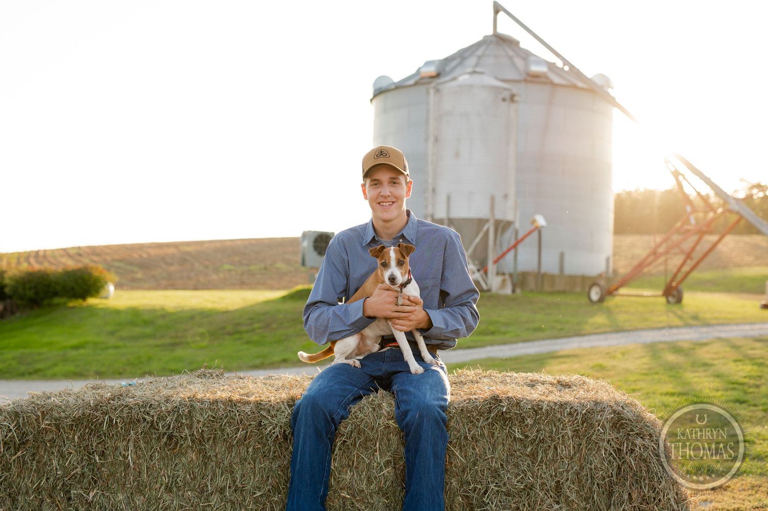 Person and dog pose in front of white grain silo during sunset on a farm.