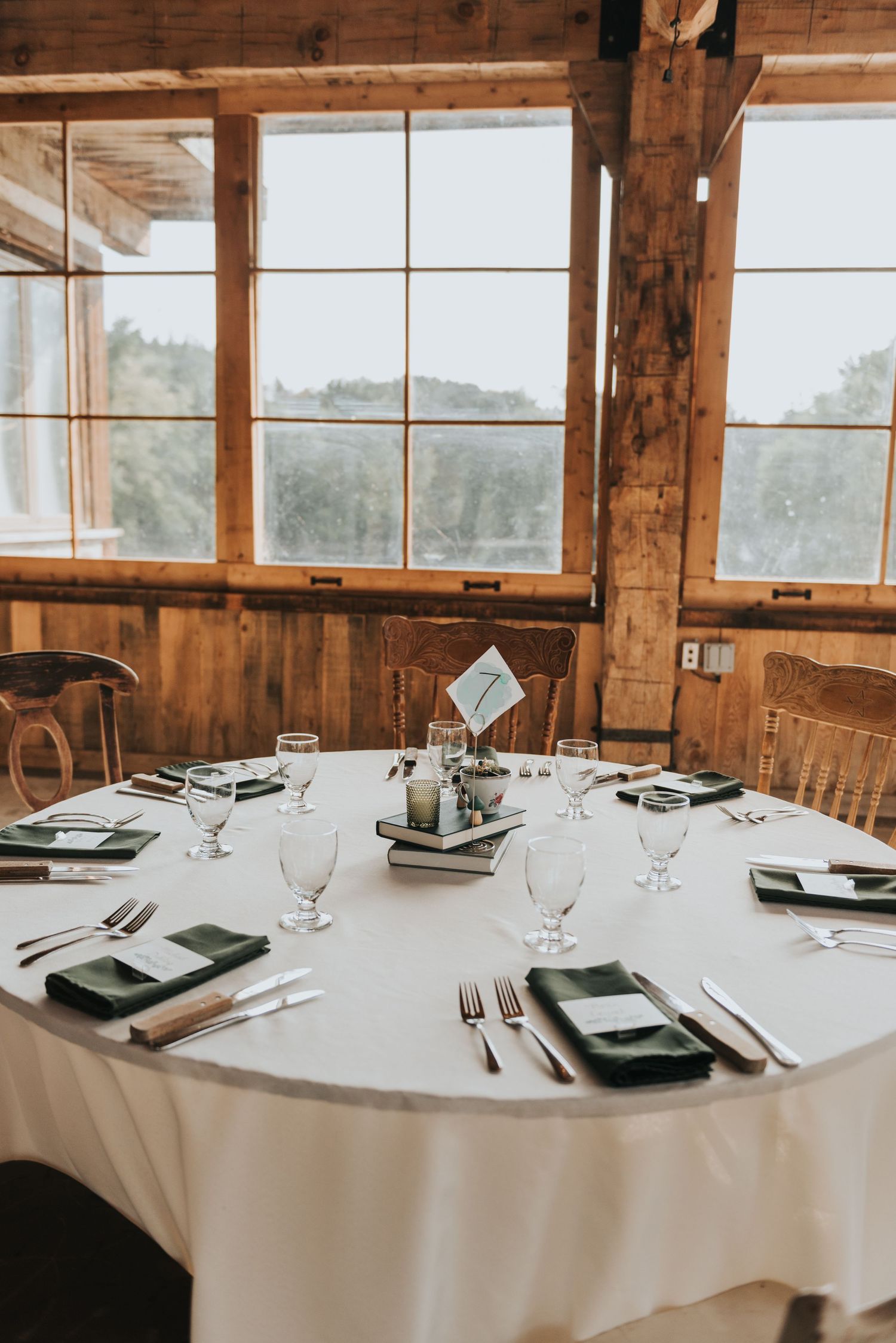 Elegant table setting in rustic wooden venue with white tablecloth and large windows overlooking scenic landscape.