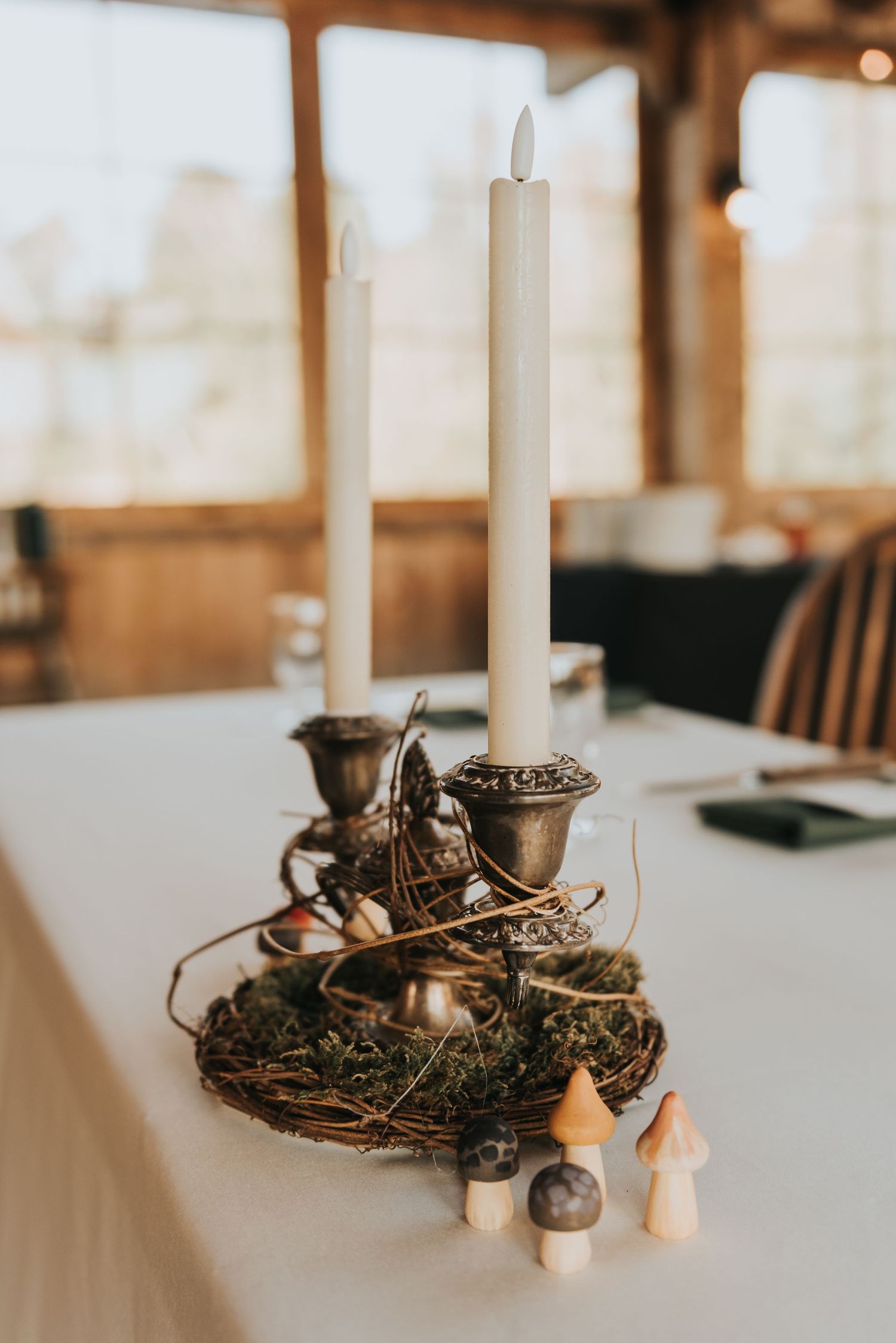 Elegant candlestick centerpiece with white tapers in silver holders surrounded by moss, twine and small wooden decorations.