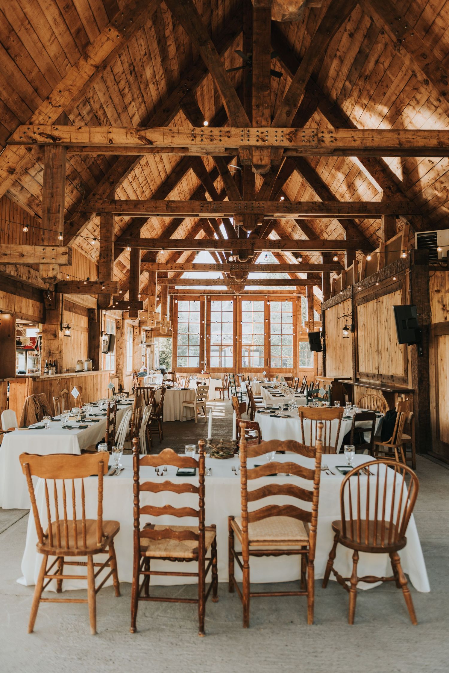 Rustic barn wedding venue interior with exposed wooden beams and mismatched wooden chairs at white-clothed tables.