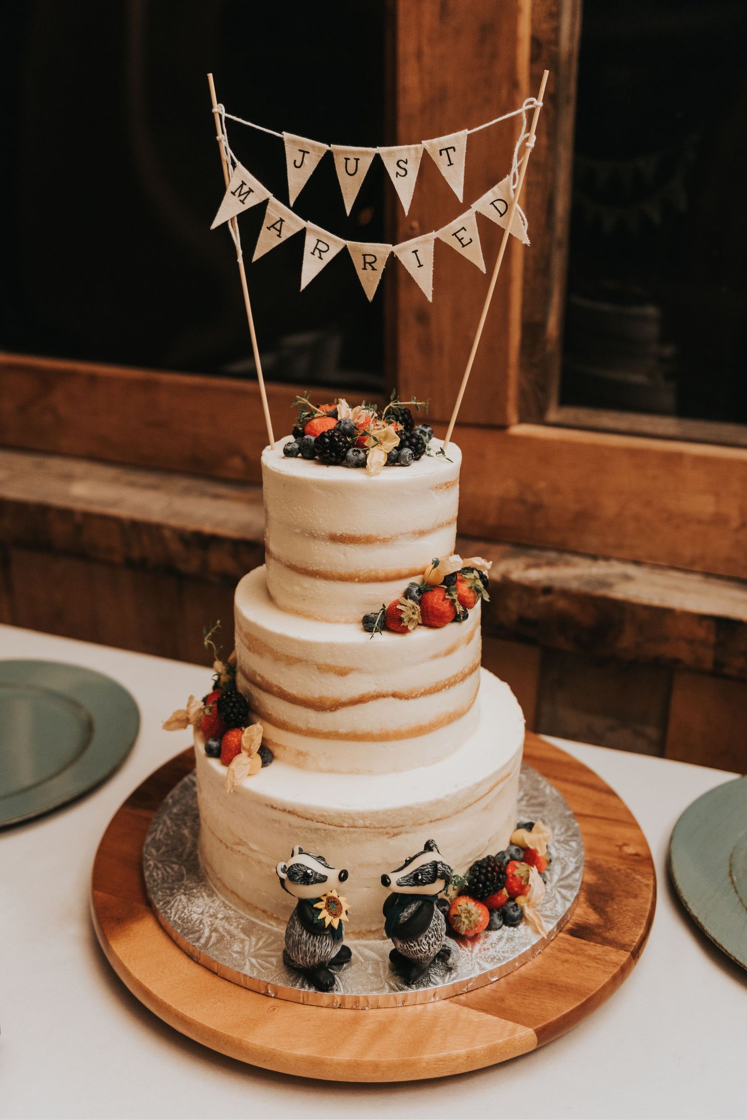 A three-tiered semi-naked wedding cake decorated with fresh berries and a 'Just Married' banner topper on a wooden board.
