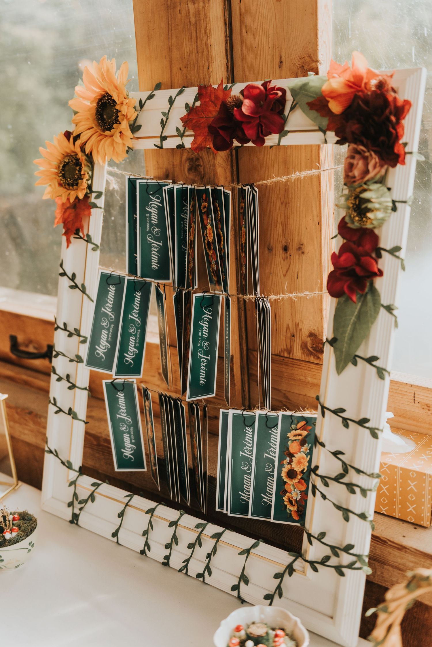 A rustic wedding seating chart display with flowers and greenery featuring escort cards on strings in a wooden frame.