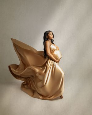 Maternity portrait in flowing gold silk dress against light neutral background with dramatic fabric movement.