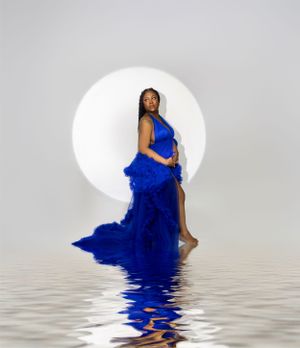 A striking artistic portrait in royal blue dress against a white circular backdrop with water reflection below.