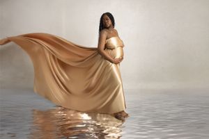 Pregnant woman wearing flowing beige gown standing in reflective water during maternity photoshoot.