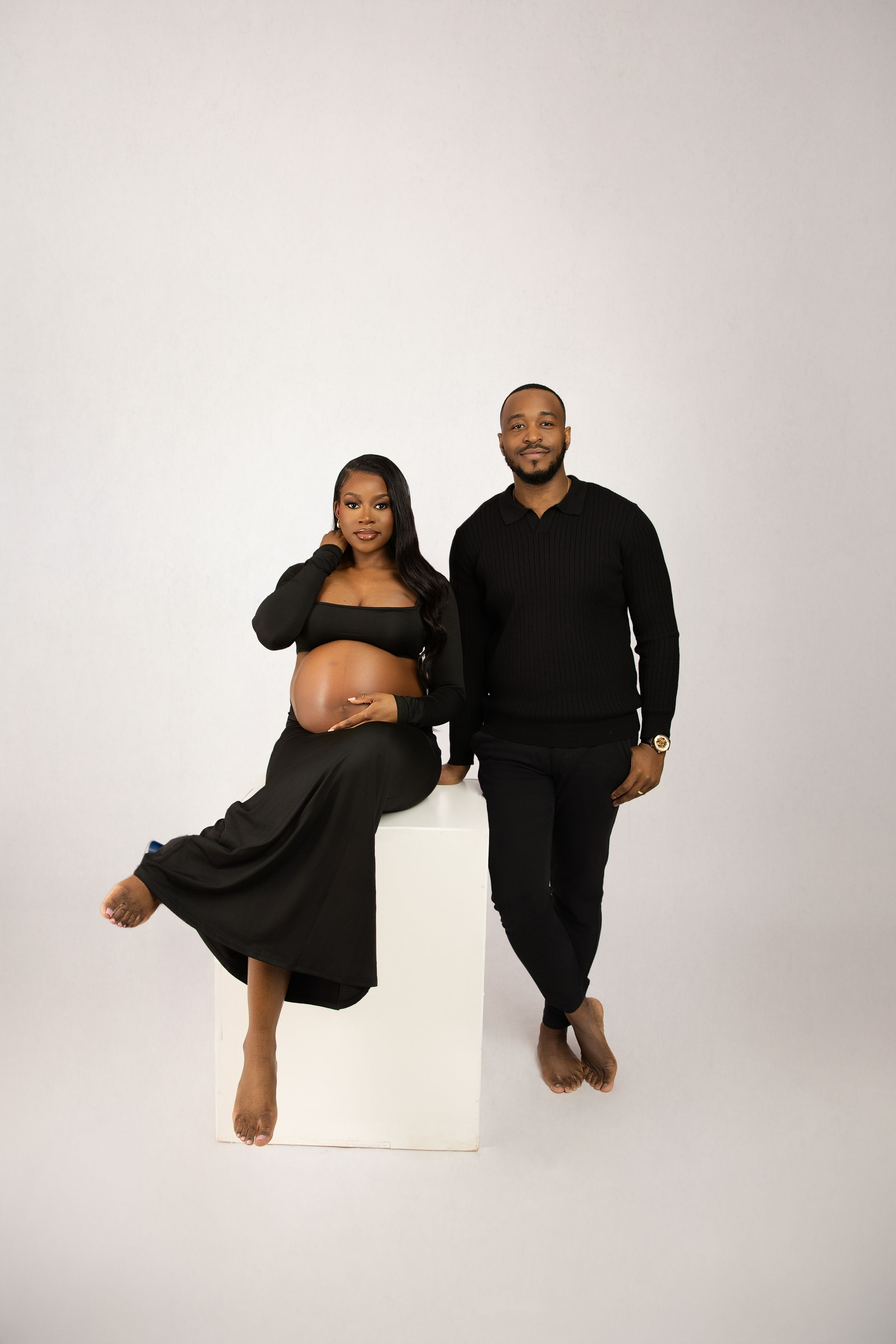A couple in matching black outfits poses together against a white backdrop during a studio photoshoot.