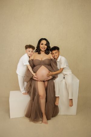 A maternity portrait featuring a woman in a brown tulle gown with two children in white outfits sitting on blocks against beige.