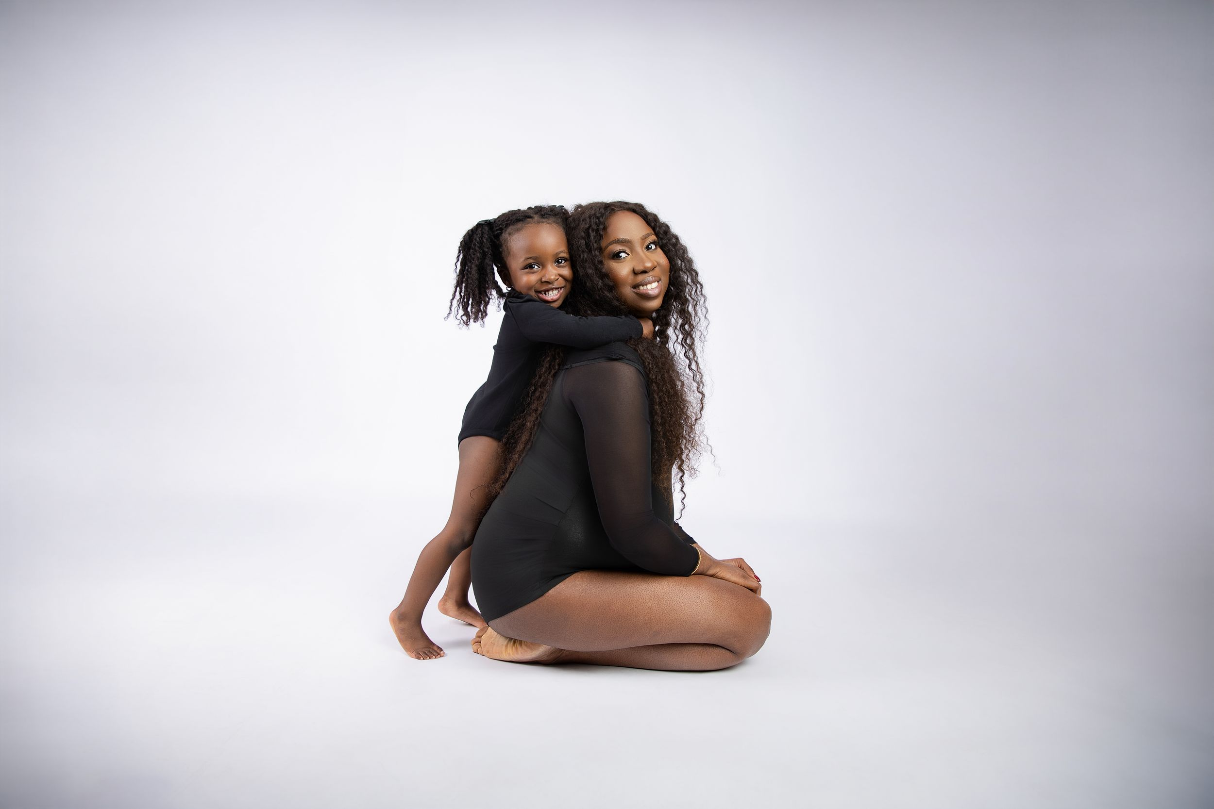 Two black friends sitting back to back and smiling joyfully in a studio setting against a soft gray background.