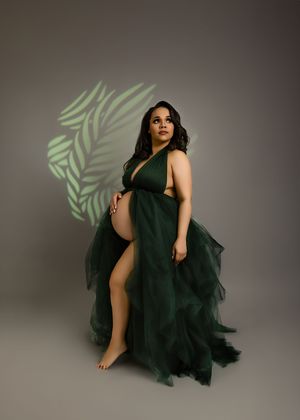 Artistic maternity photo with a flowy emerald green tulle gown and projected palm leaf shadow against grey backdrop.