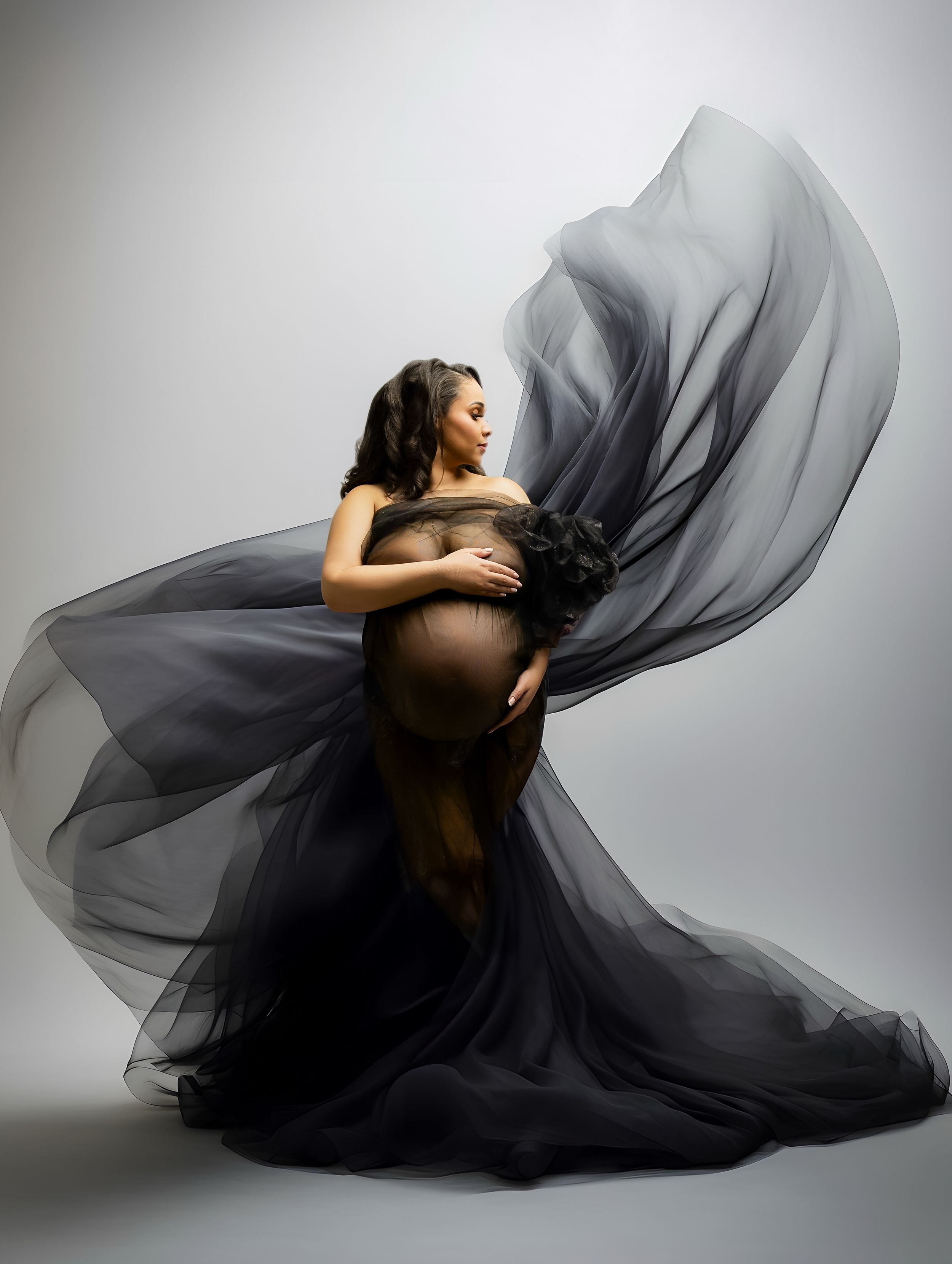 Artistic maternity photo with flowing black tulle fabric dramatically billowing against light gray studio background.
