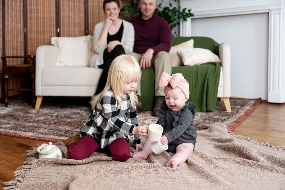 Family of four relaxing at home, two parents watching their young daughters play on a blanket.