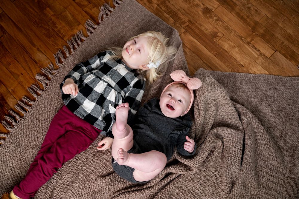 Top view of two young sisters lying on a brown blanket, baby laughing with toddler beside her.