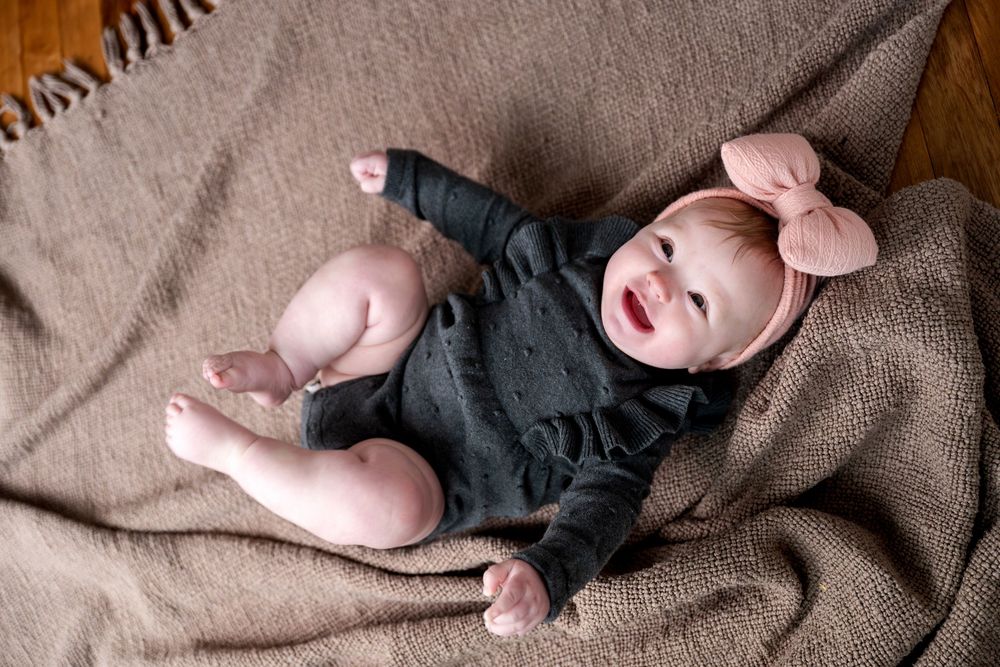 Adorable baby girl in dark romper and pink bow headband lying on a brown textured blanket.