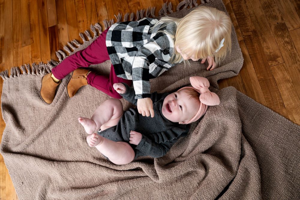 Overhead view of blonde toddler leaning over laughing baby sister on a brown blanket floor.