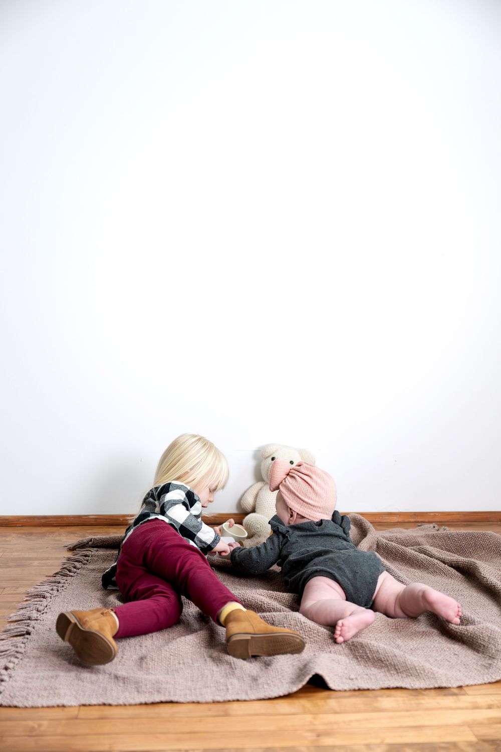 Two young sisters sitting on a brown blanket sharing a stuffed bunny toy against a white wall.