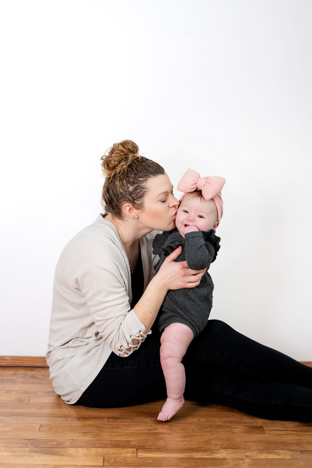 A mother tenderly kisses her smiling baby girl wearing a pink bow headband while sitting on hardwood floor.