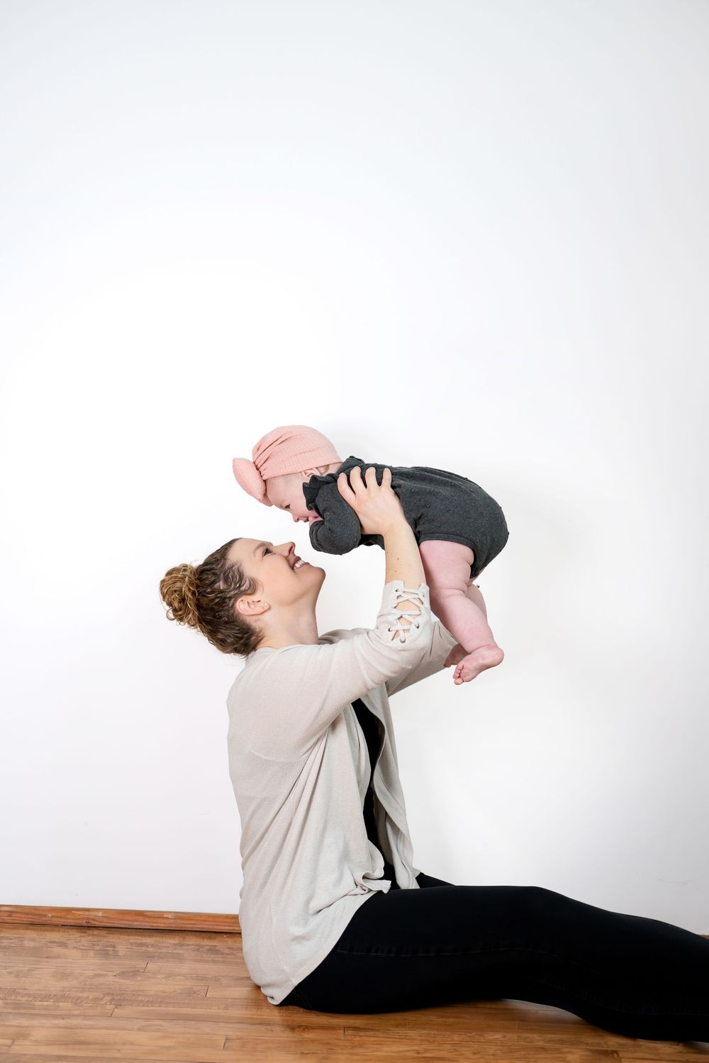 A joyful mother lifts her baby girl up in the air while sitting on a hardwood floor against a white wall.