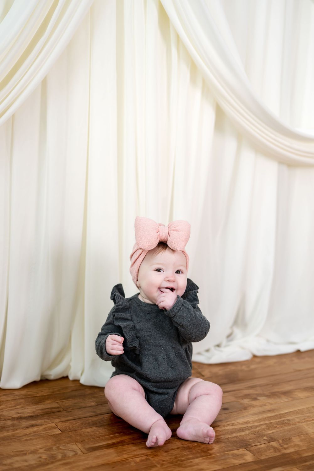 Adorable baby girl in gray ruffle outfit and pink bow headband sitting before white curtains.