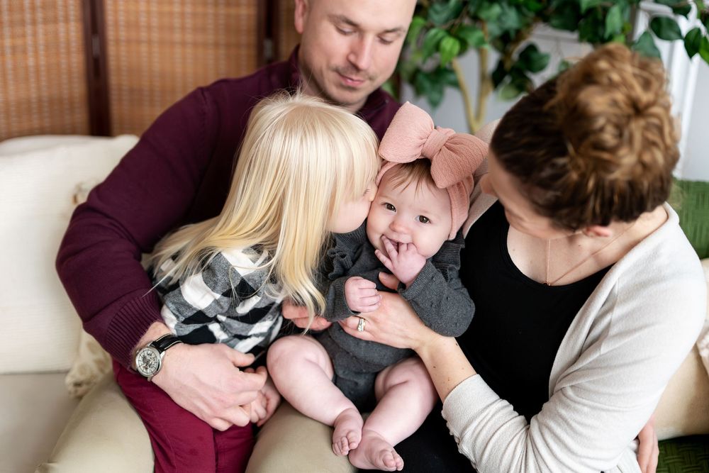 Parents and toddler girl lovingly interacting with baby dressed in gray on white couch.