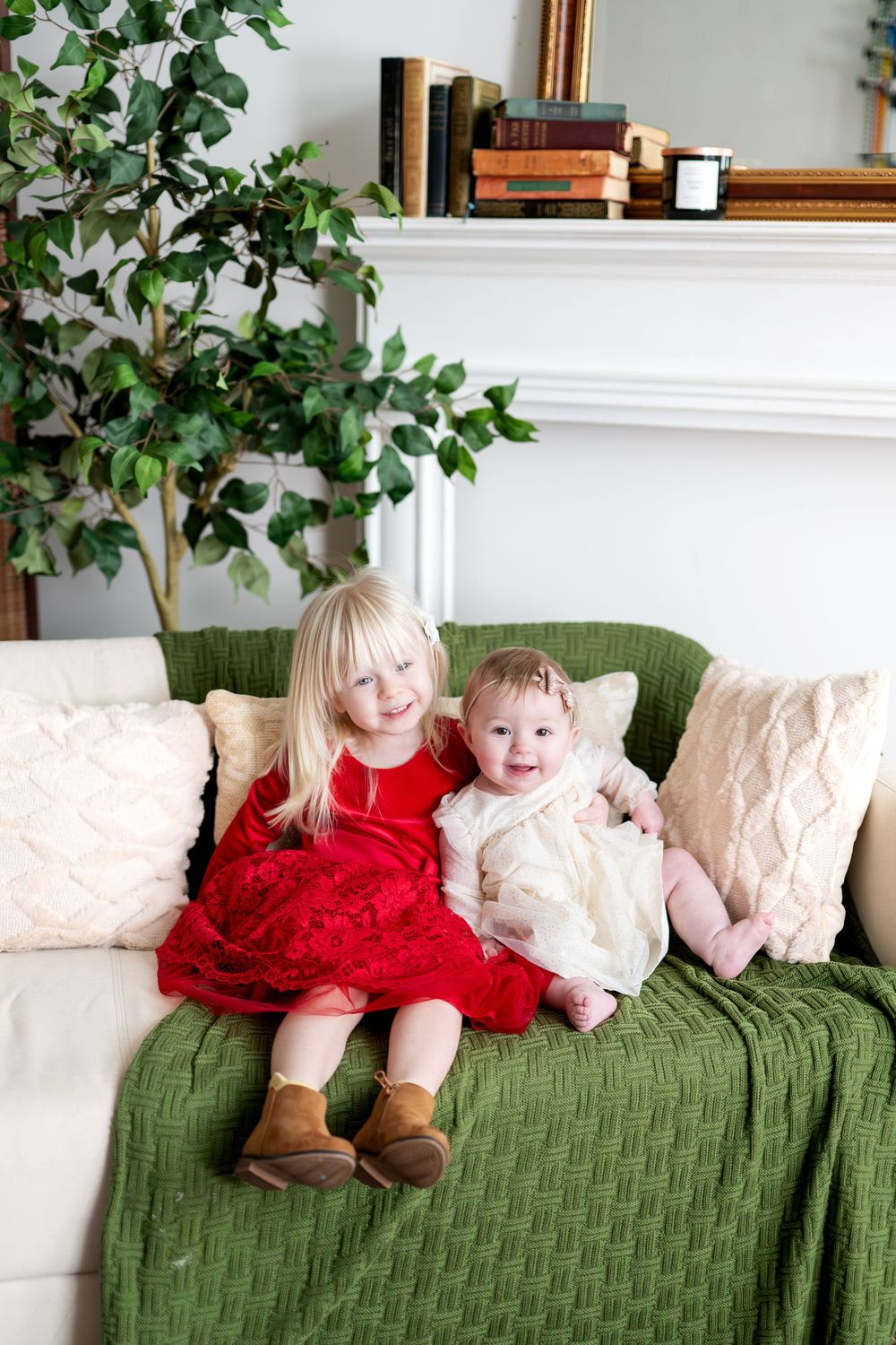 Two young sisters sitting together on a couch with green blanket, older girl in red dress hugging baby in white dress.