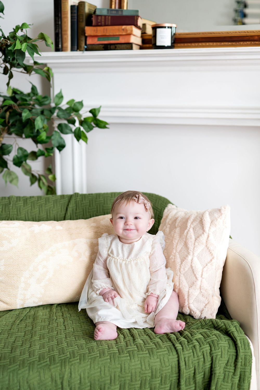 Sweet baby girl in cream white dress sitting on green textured blanket on couch smiling at camera.