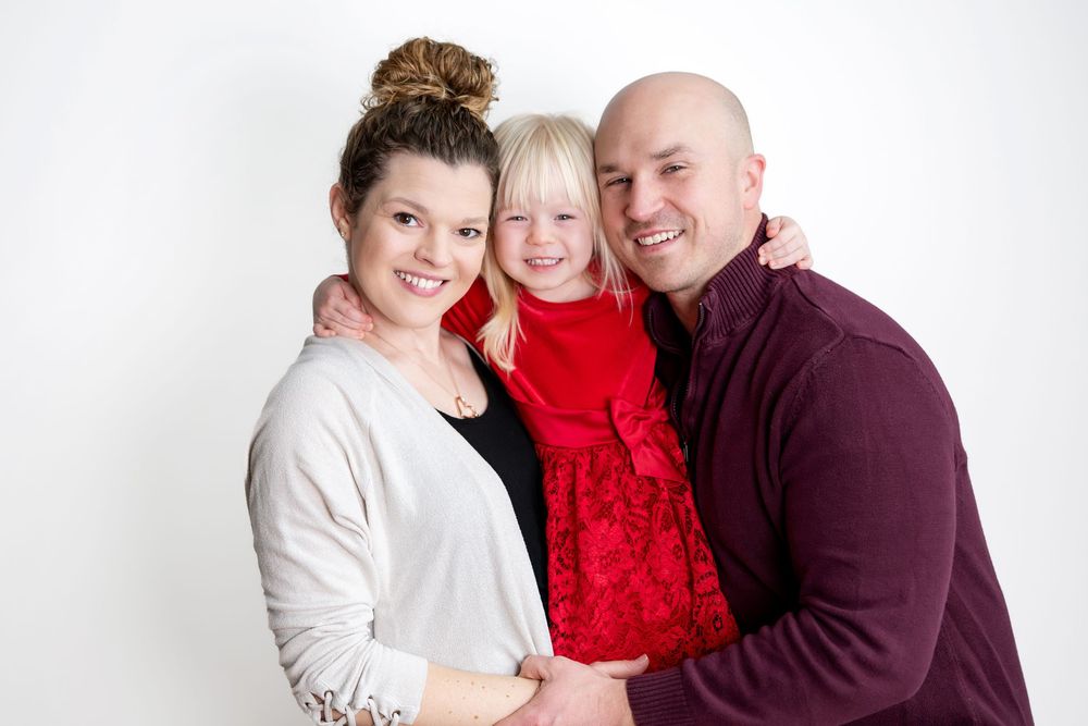 Happy family of three posing together, parents smiling with young blonde girl in red dress against white background.