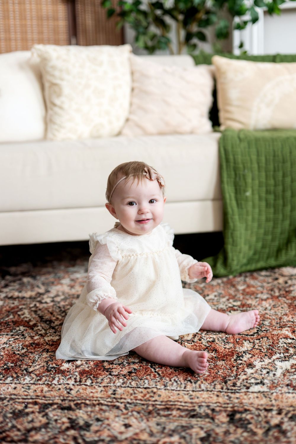 Smiling baby girl in white dress sitting on ornate rug in cozy living room.