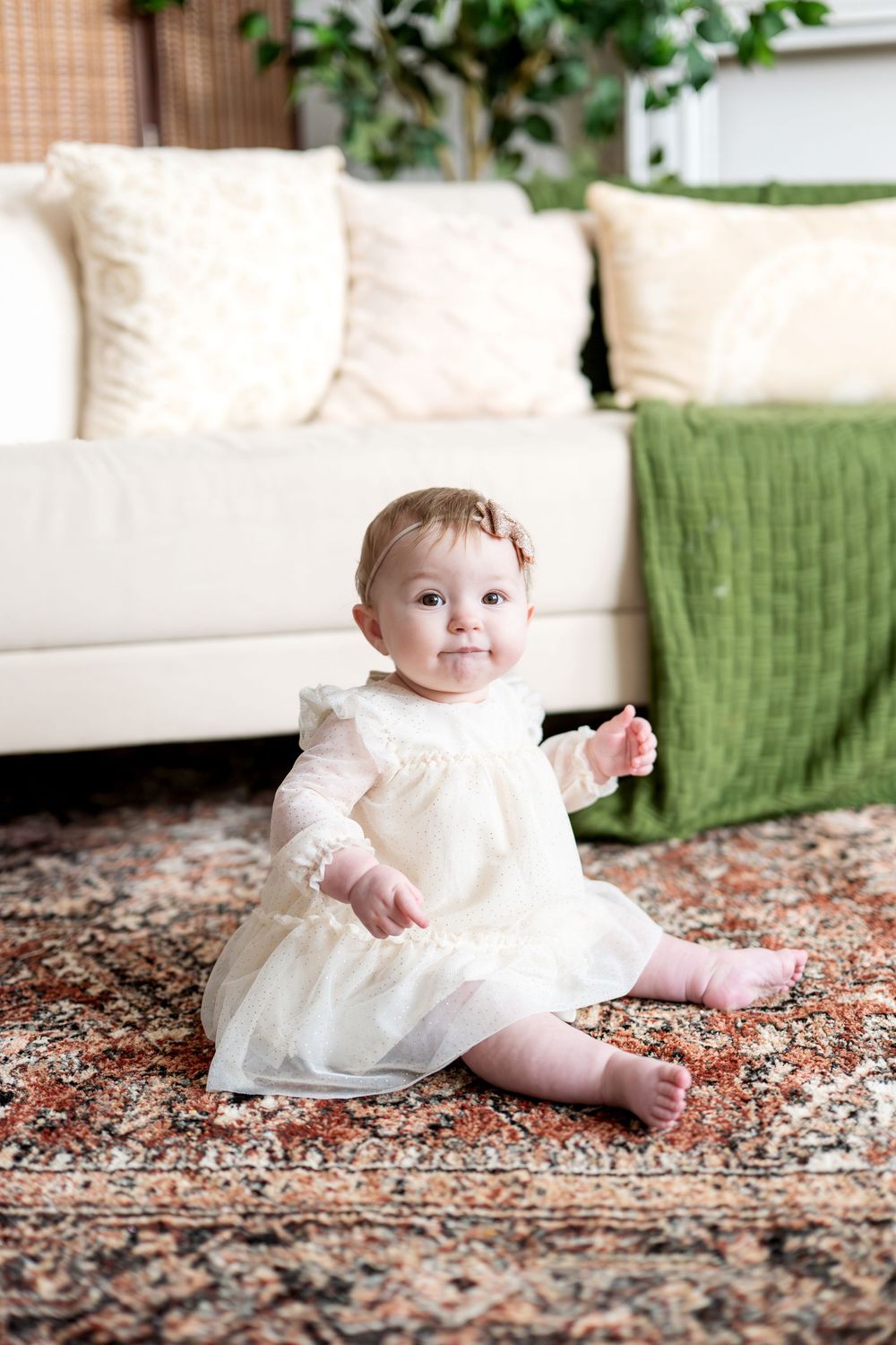 Adorable baby girl in cream dress sitting on patterned rug near white sofa.