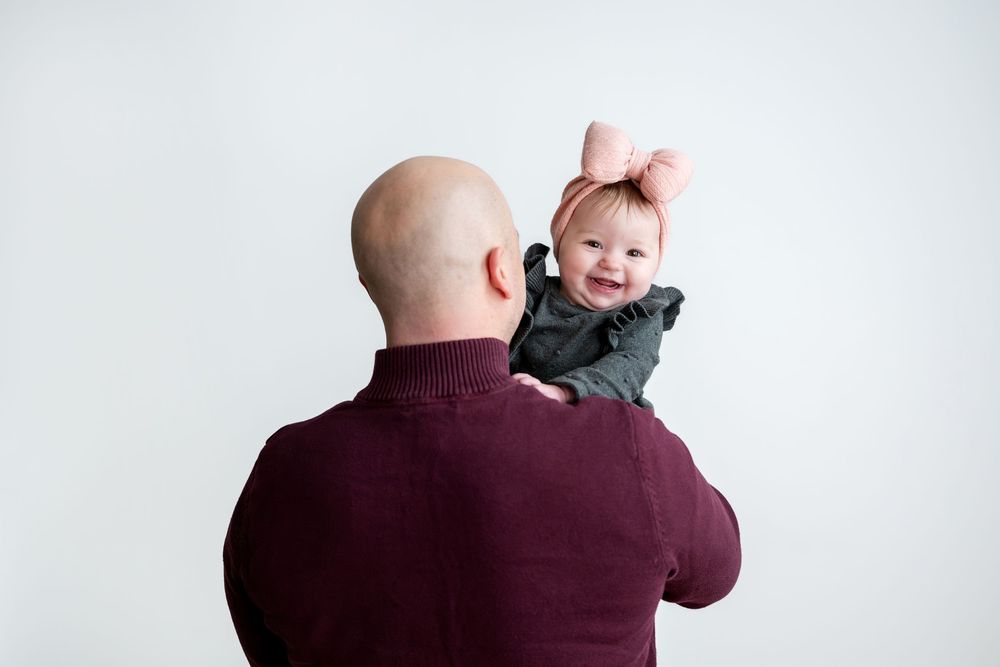 Bald father lifting smiling baby girl with pink bow against white background.