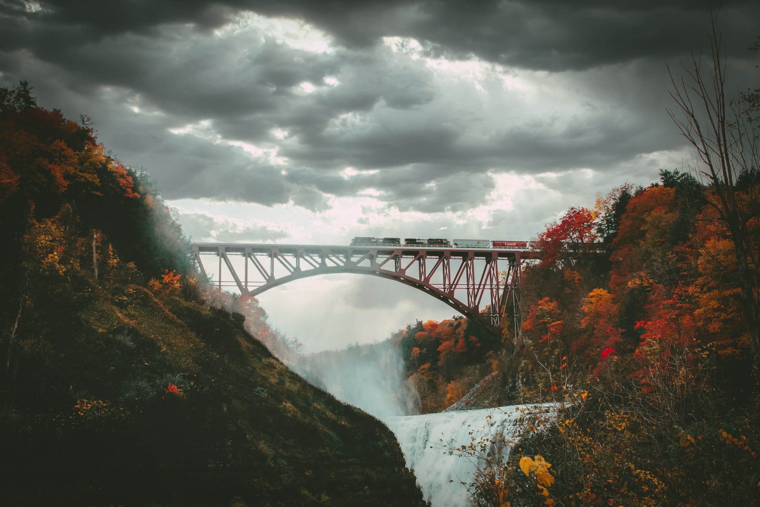 Dramatic storm clouds gather over a steel arch bridge surrounded by autumn foliage.