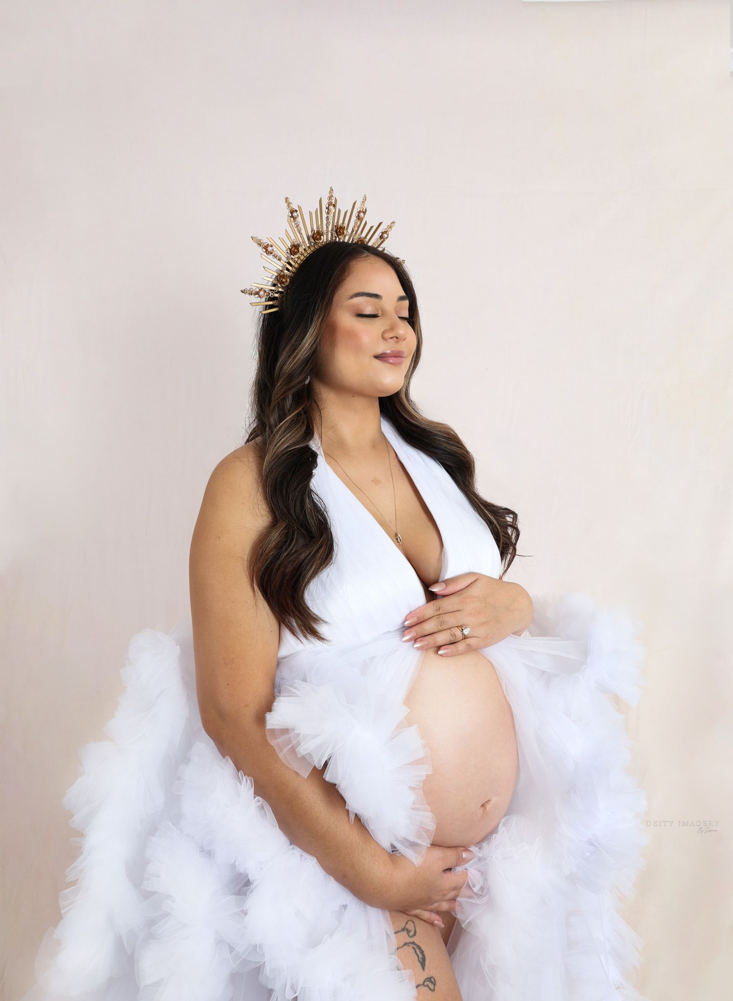 Ethereal maternity photo featuring a white tulle gown and gold crown against a light backdrop.