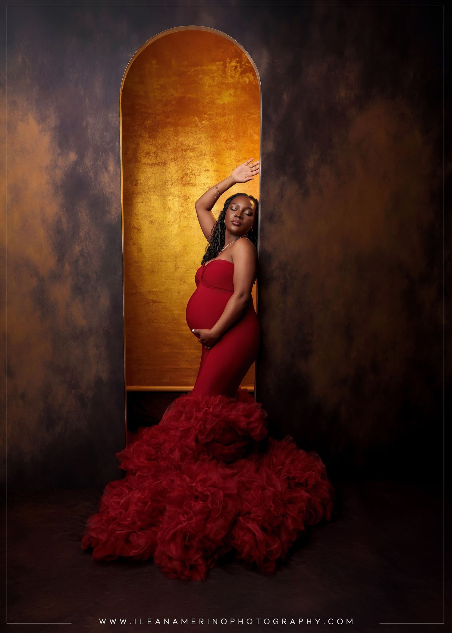 Pregnant woman in red ruffled gown posing in golden arch on dark textured background.