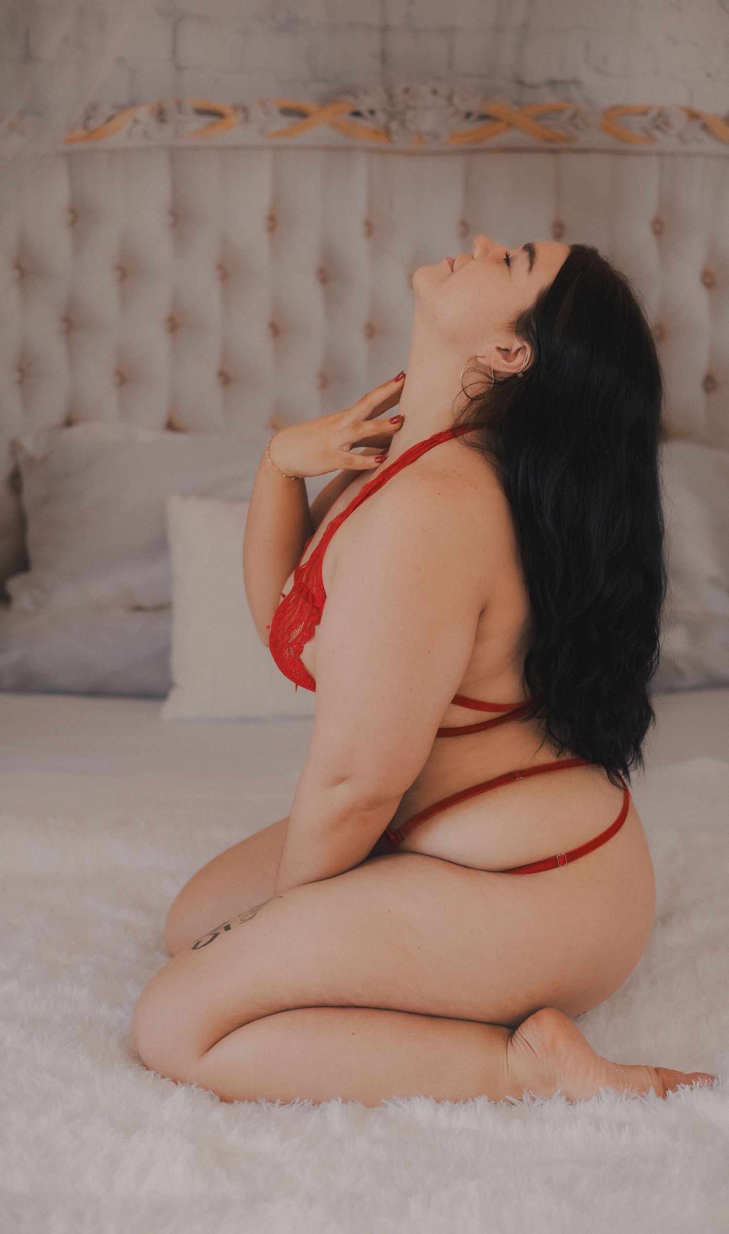 A woman in red lingerie poses on a white bed with tufted headboard in a boudoir photography session.