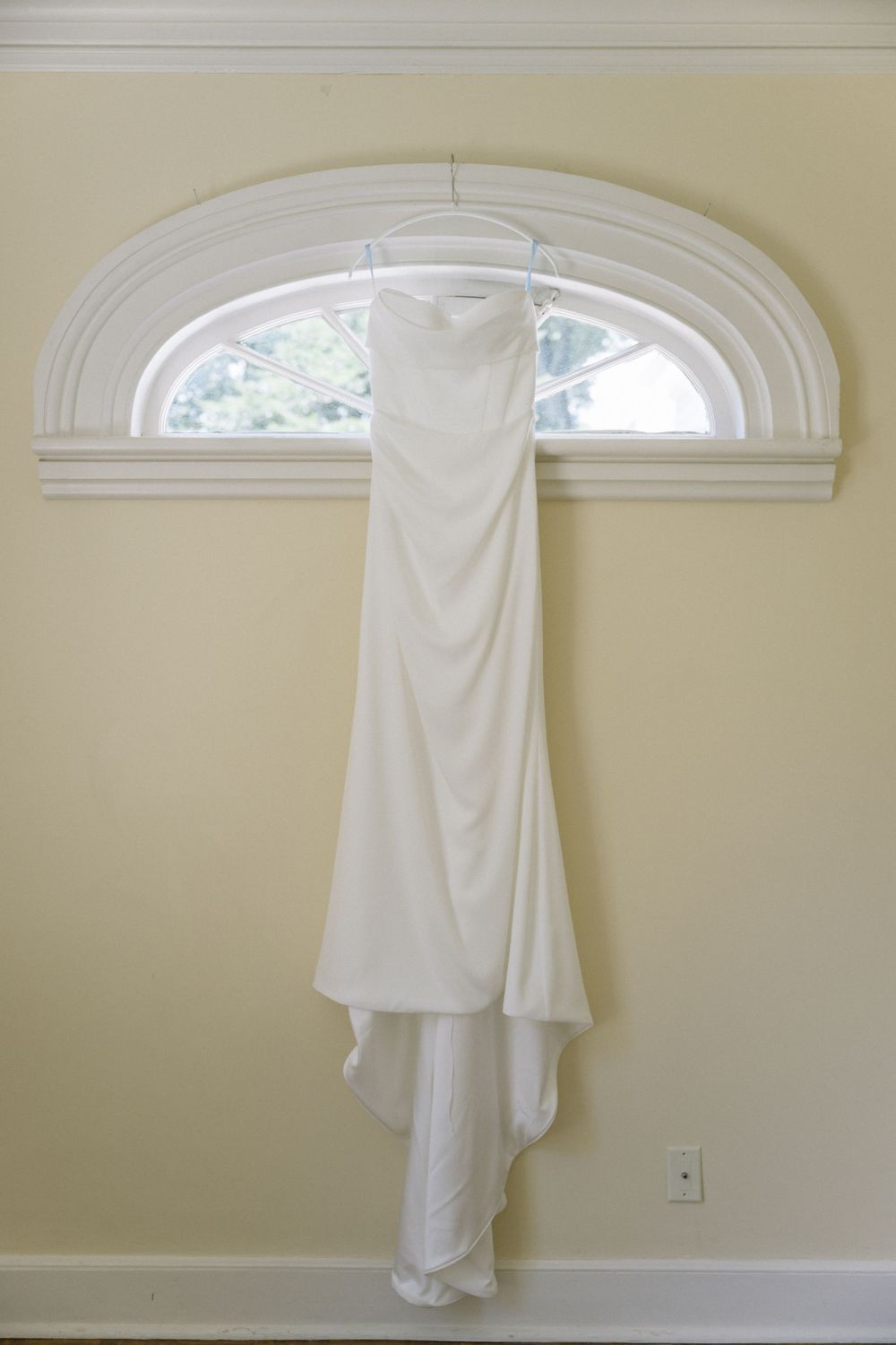 Elegant white wedding gown hanging from arched window frame against cream colored wall.
