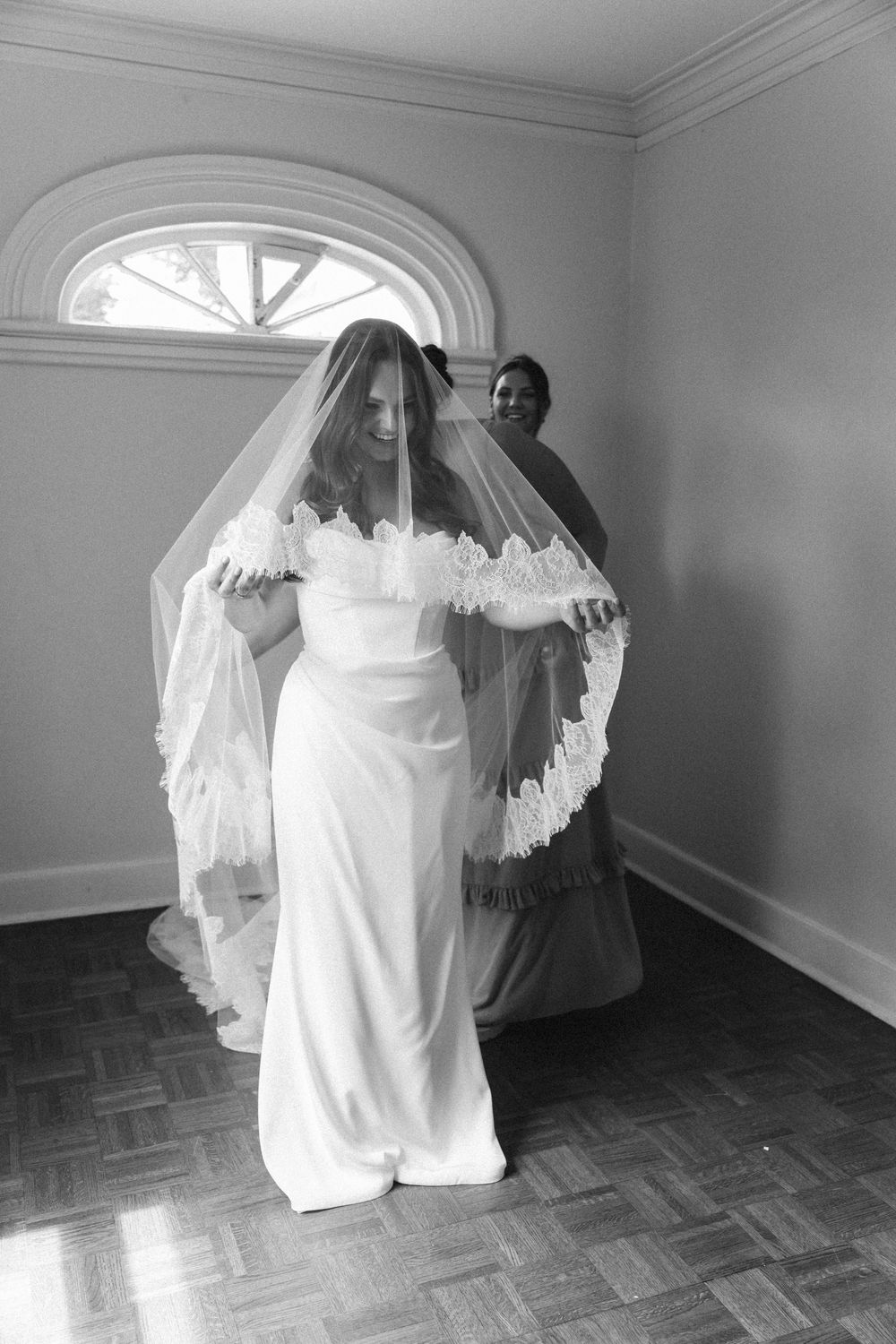Classic black and white photo of wedding dress preparation in an elegant room with arched window.