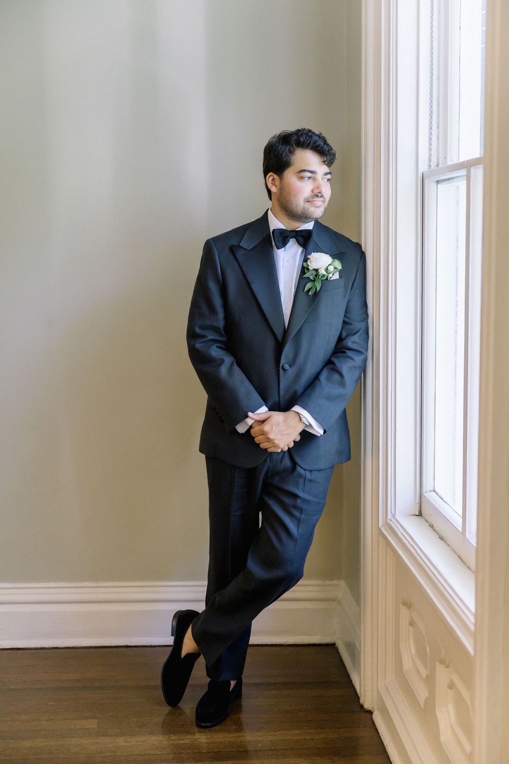 Formal portrait against white wall showing black suit and boutonniere details.