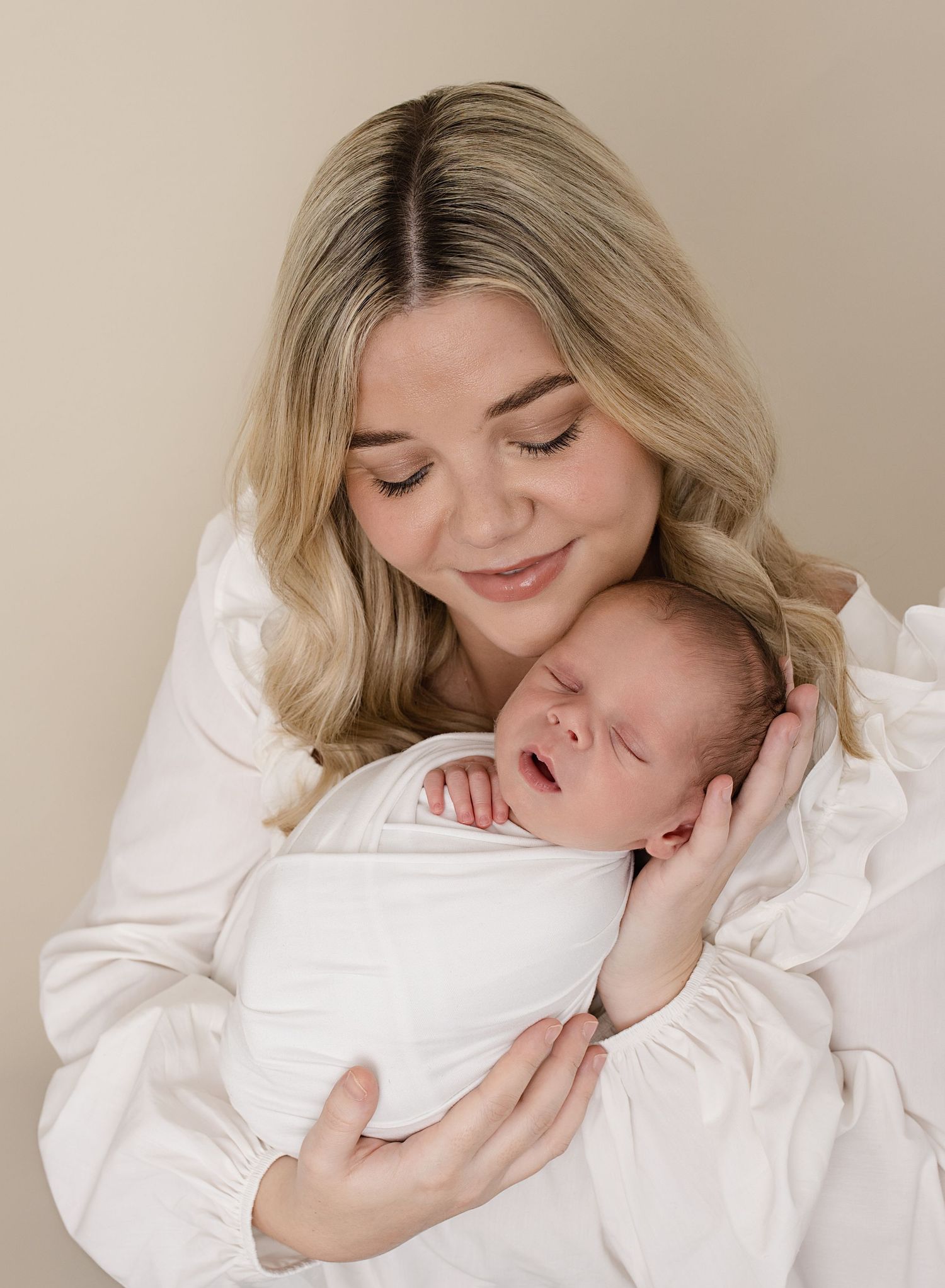 Warm image of beautiful new mum wearing white, holding her tiny newborn close and looking down on him as he sleeps during photoshoot
