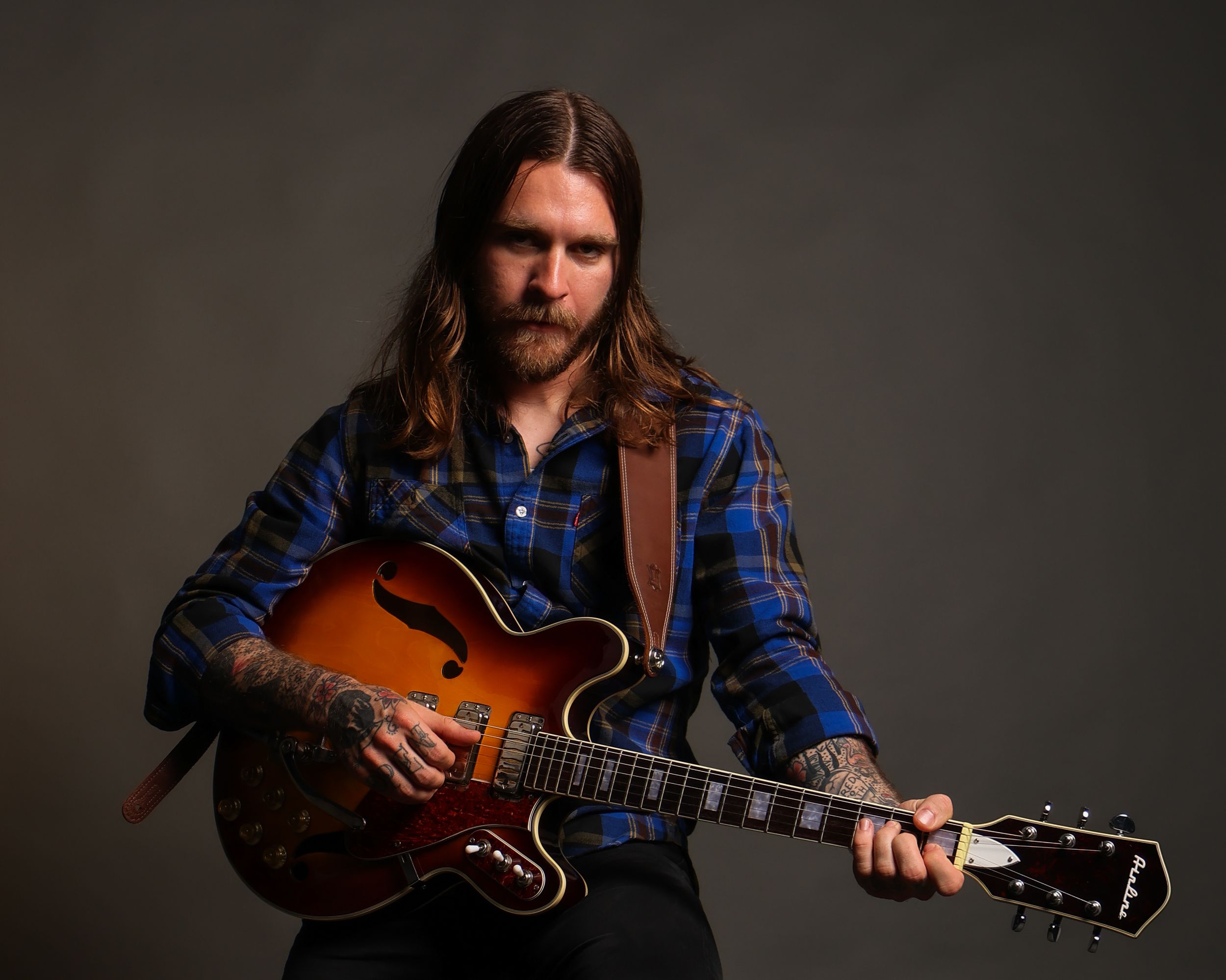 A musician in a blue plaid shirt plays an acoustic guitar in a series of moody studio portraits.