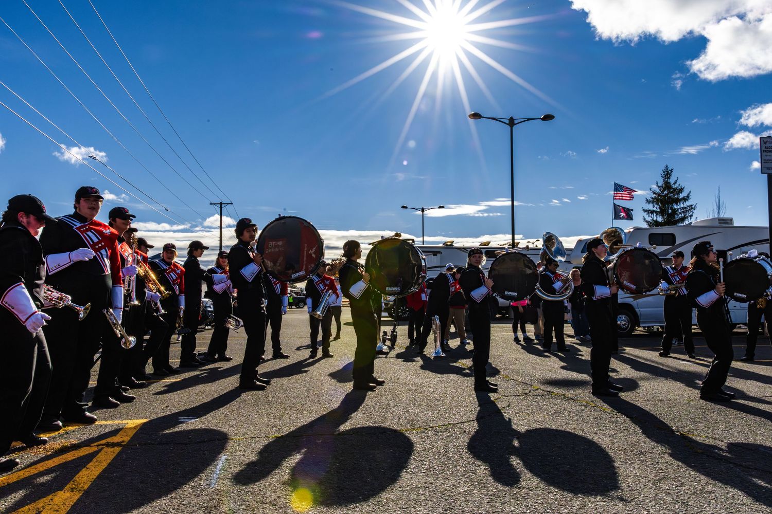 A crowd of people cast long shadows in a parkinglot during a bright sunny day with blue skies.