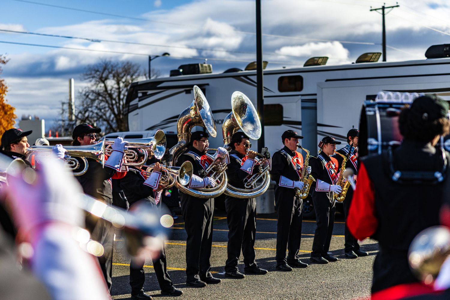 A marching band performs outdoors in formation while playing brass instruments on a sunny day with clouds overhead.