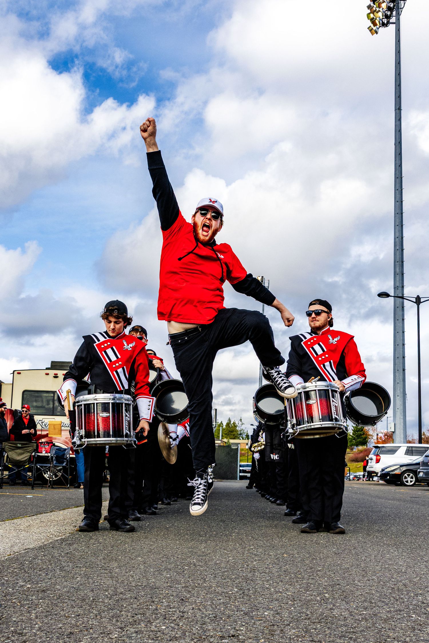 Energetic performer jumps high between marching band drummers in red and black uniforms under cloudy sky.